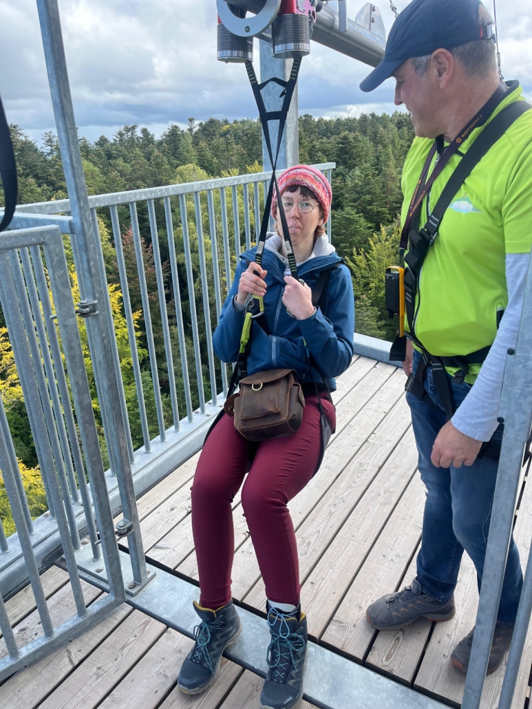 Rika ist auf einer Aussichtsplattform fest im Sitz der Attraktion "Flyline" angeschnallt, neben ihr steht ein Mitarbeiter in einem hellgrünen T-Shirt.
